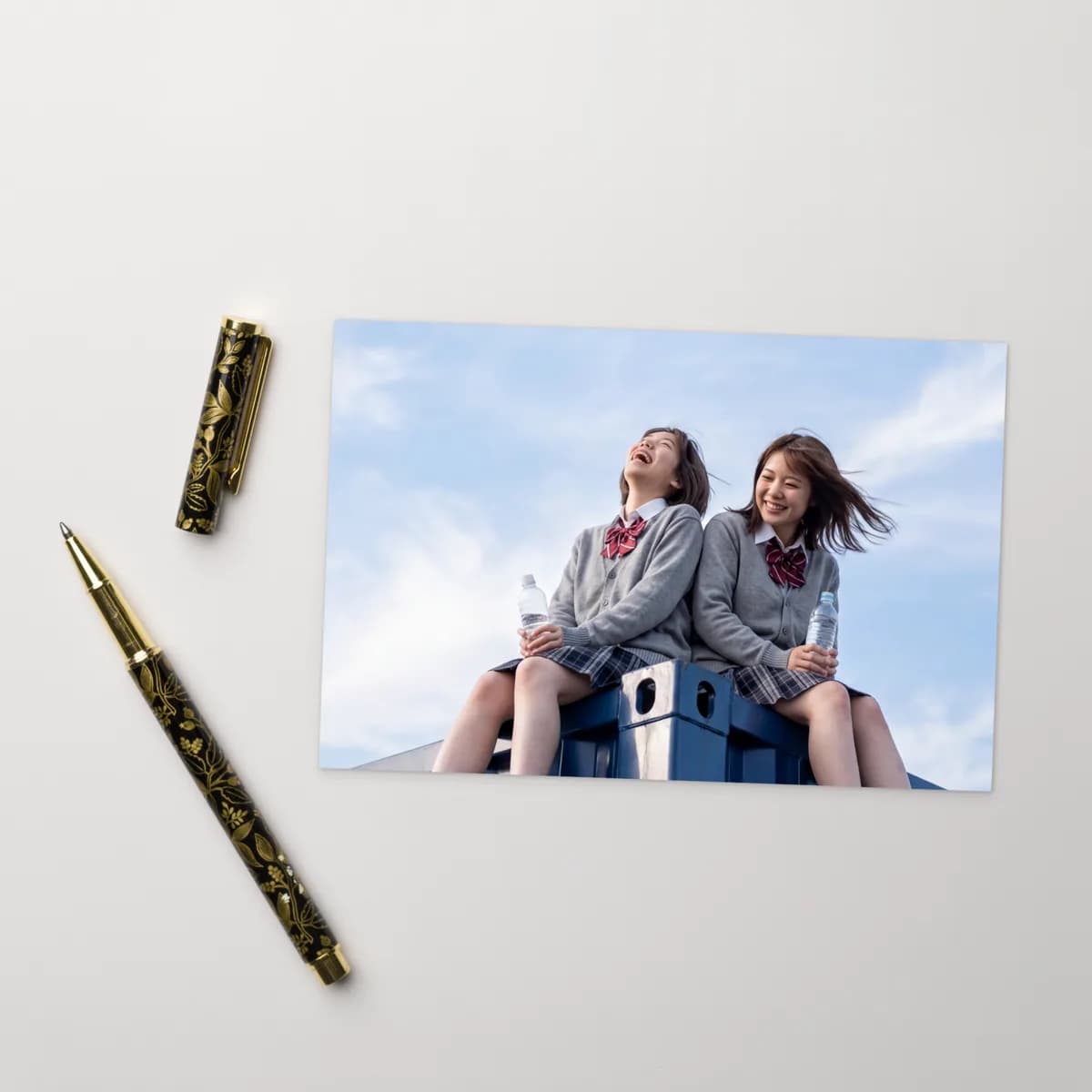 The postcard on a white desk beside a gold rollerball pen, showing two friends in school uniforms laughing under a blue sky